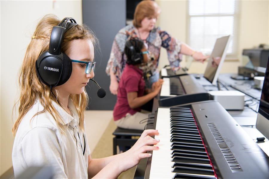 A student plays a piano while wearing a headset and a teacher assists another student.